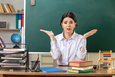 concerned-spreading-hands-young-female-teacher-sitting-at-table-with-school-tools-in-classroom.jpg